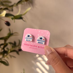 Puede incluir: Un par de pendientes de arcilla polim&eacute;rica blanca y azul con forma del personaje R2-D2 de Star Wars. Los pendientes est&aacute;n en una tarjeta rosa con el texto "SWEET LOVE CLAY" y el nombre de usuario de las redes sociales "@sweetloveclay".