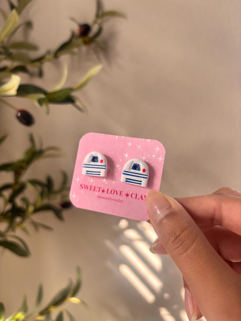 Puede incluir: Un par de pendientes de arcilla polim&eacute;rica blanca y azul con forma del personaje R2-D2 de Star Wars. Los pendientes est&aacute;n en una tarjeta rosa con el texto "SWEET LOVE CLAY" y el nombre de usuario de las redes sociales "@sweetloveclay".