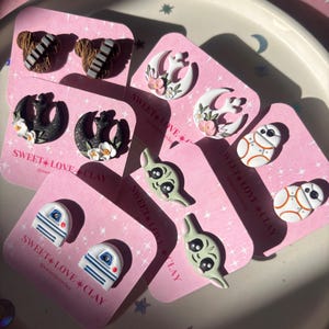 Puede incluir: Un conjunto de 10 pendientes hechos a mano de arcilla polim&eacute;rica con personajes y s&iacute;mbolos de la franquicia Star Wars. Los pendientes se muestran en tarjetas rosas con el texto "SWEET LOVE CLAY" y el nombre de usuario de las redes sociales "@sweetloveclay".