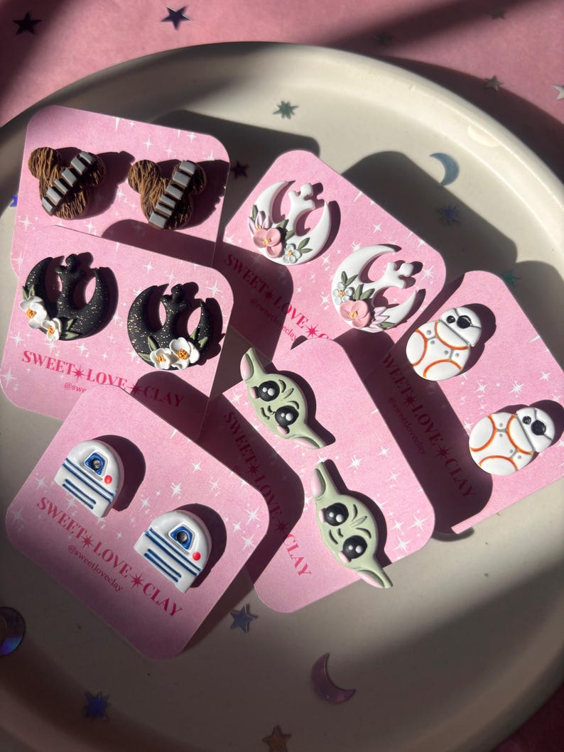 Puede incluir: Un conjunto de 10 pendientes hechos a mano de arcilla polim&eacute;rica con personajes y s&iacute;mbolos de la franquicia Star Wars. Los pendientes se muestran en tarjetas rosas con el texto "SWEET LOVE CLAY" y el nombre de usuario de las redes sociales "@sweetloveclay".