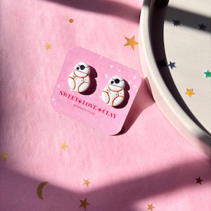 Puede incluir: Un par de pendientes de arcilla polim&eacute;rica blanca y naranja con forma del personaje de Star Wars BB-8. Los pendientes est&aacute;n en una tarjeta blanca con el texto "SWEET LOVE CLAY @sweetloveclay".