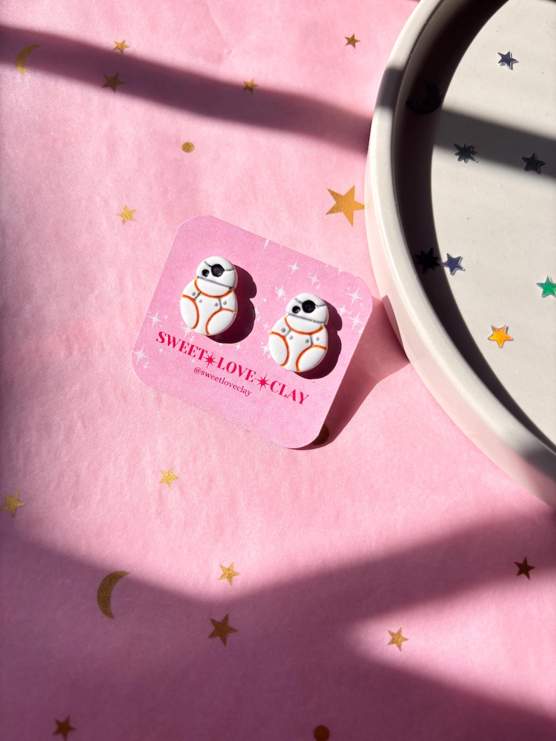 Puede incluir: Un par de pendientes de arcilla polim&eacute;rica blanca y naranja con forma del personaje de Star Wars BB-8. Los pendientes est&aacute;n en una tarjeta blanca con el texto "SWEET LOVE CLAY @sweetloveclay".