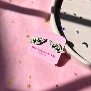 Puede incluir: Un par de pendientes de arcilla verde y blanca con un personaje de dibujos animados con ojos grandes. Los pendientes est&aacute;n en una tarjeta blanca con el texto "SWEET LOVE CLAY" y el nombre de usuario de las redes sociales "@sweetloveclay".