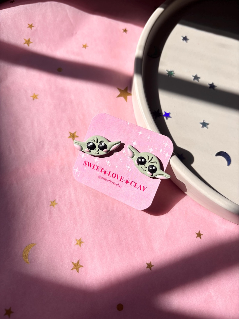 Puede incluir: Un par de pendientes de arcilla verde y blanca con un personaje de dibujos animados con ojos grandes. Los pendientes est&aacute;n en una tarjeta blanca con el texto "SWEET LOVE CLAY" y el nombre de usuario de las redes sociales "@sweetloveclay".