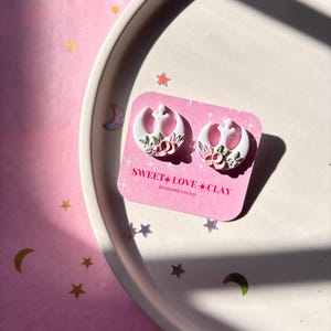 Puede incluir: Un par de pendientes blancos con el s&iacute;mbolo de la Alianza Rebelde con detalles florales rosas y blancos. Los pendientes se muestran en un plato blanco con fondo rosa. El texto "SWEET*LOVE*CLAY @sweetloveclay" est&aacute; impreso en una tarjeta rosa.
