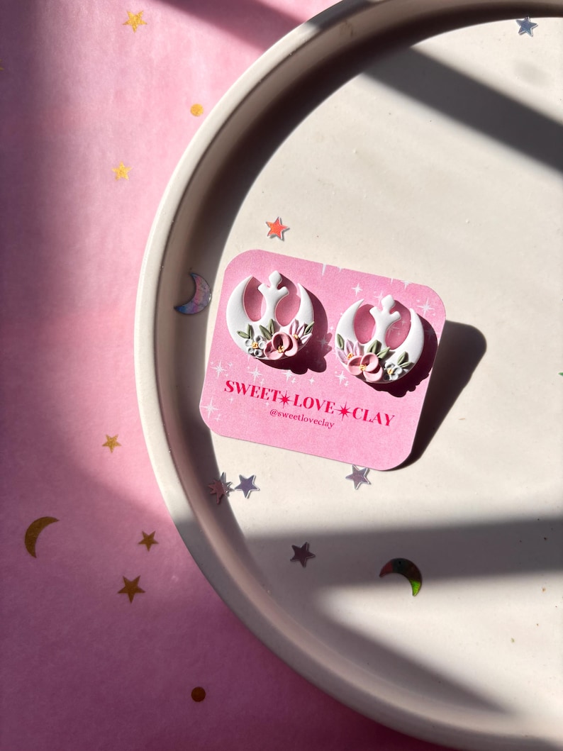 Puede incluir: Un par de pendientes blancos con el s&iacute;mbolo de la Alianza Rebelde con detalles florales rosas y blancos. Los pendientes se muestran en un plato blanco con fondo rosa. El texto "SWEET*LOVE*CLAY @sweetloveclay" est&aacute; impreso en una tarjeta rosa.