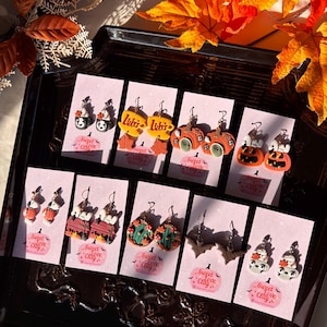 May include: A selection of Halloween-themed earrings presented on pink cards. The earrings showcase diverse designs, such as pumpkins, bats, and cartoon characters. The cards bear the text "Sweet Love Clay" and are arranged on a black tray.