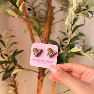 Puede incluir: Un par de pendientes de coraz&oacute;n marr&oacute;n y blanco con un patr&oacute;n a rayas. Los pendientes est&aacute;n en una tarjeta rosa con el texto "SWEET LOVE CLAY @sweetloveclay".