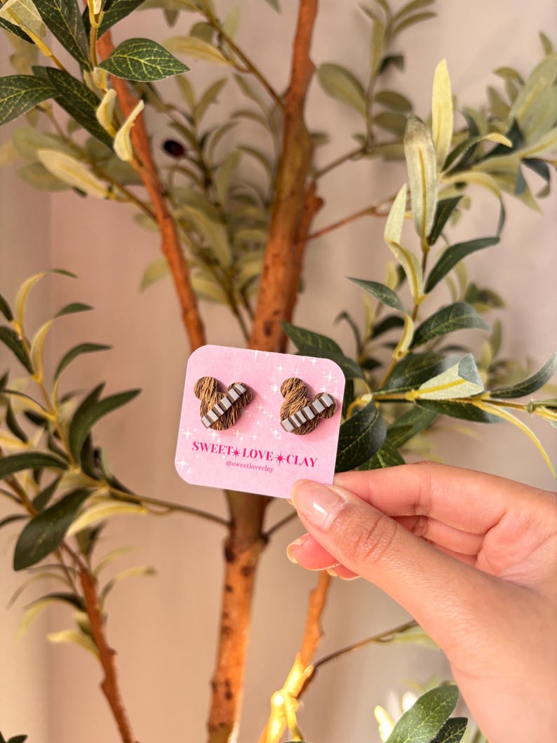Puede incluir: Un par de pendientes de coraz&oacute;n marr&oacute;n y blanco con un patr&oacute;n a rayas. Los pendientes est&aacute;n en una tarjeta rosa con el texto "SWEET LOVE CLAY @sweetloveclay".