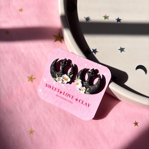 Puede incluir: Un par de pendientes de media luna negra con detalles de flores blancas. Los pendientes est&aacute;n sobre un fondo rosa con estrellas doradas. El texto "SWEET LOVE CLAY" y "@sweetloveclay" est&aacute; en la tarjeta.