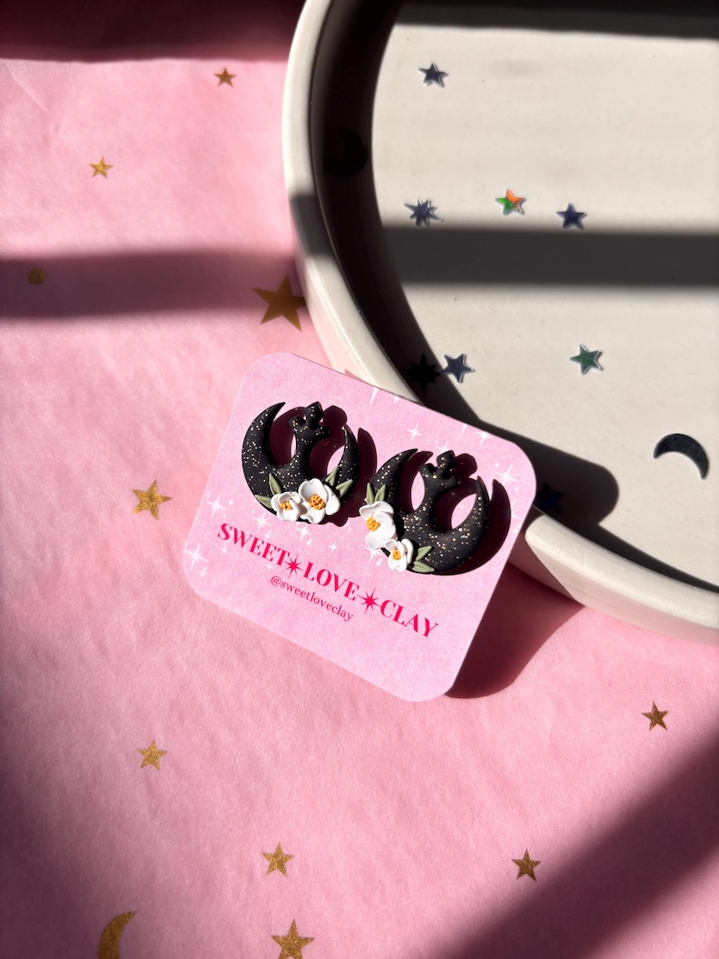 Puede incluir: Un par de pendientes de media luna negra con detalles de flores blancas. Los pendientes est&aacute;n sobre un fondo rosa con estrellas doradas. El texto "SWEET LOVE CLAY" y "@sweetloveclay" est&aacute; en la tarjeta.