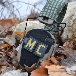 Peut inclure: Casque tactique beige et noir avec les lettres "MC" en jaune sur un patch noir. Le casque repose sur un lit de feuilles mortes. Le bandeau est recouvert d'un motif &agrave; carreaux vert et beige.
