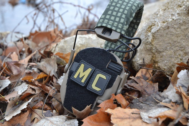 Peut inclure: Casque tactique beige et noir avec les lettres "MC" en jaune sur un patch noir. Le casque repose sur un lit de feuilles mortes. Le bandeau est recouvert d'un motif &agrave; carreaux vert et beige.