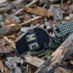 Peut inclure: Un casque tactique noir et beige, pos&eacute; sur un lit de feuilles et de bois. Le casque porte les lettres "MC" dor&eacute;es sur un patch noir. Les &eacute;couteurs sont beiges avec un motif camouflage vert et beige.