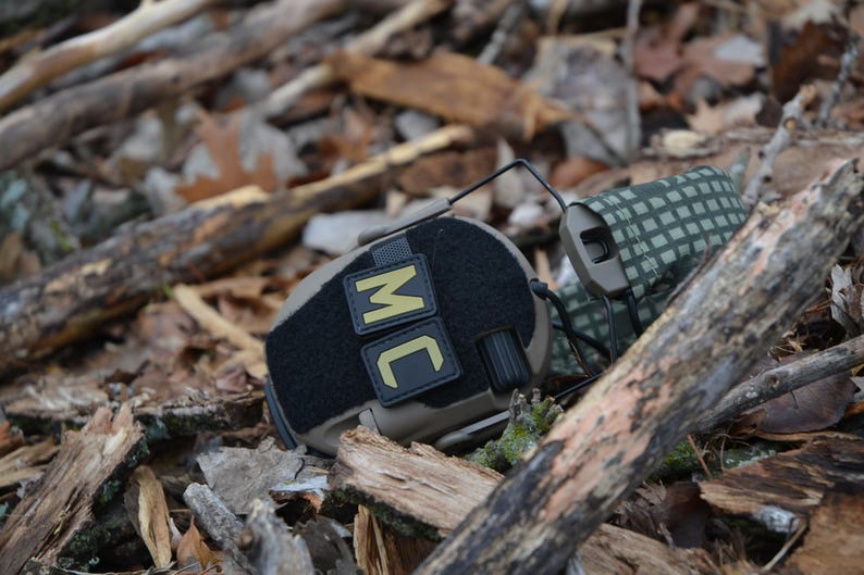 Peut inclure: Un casque tactique noir et beige, pos&eacute; sur un lit de feuilles et de bois. Le casque porte les lettres "MC" dor&eacute;es sur un patch noir. Les &eacute;couteurs sont beiges avec un motif camouflage vert et beige.
