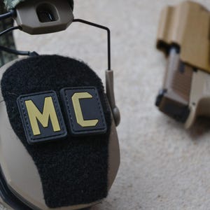 Peut inclure: Casque tactique de couleur beige avec des patchs noirs affichant "MC" en lettres jaunes. Un pistolet beige est dans un étui en arrière-plan. Le casque a une surface noire texturée et est conçu pour un usage tactique.