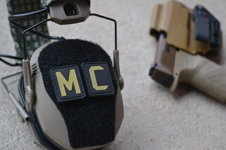 Peut inclure: Casque tactique de couleur beige avec des patchs noirs affichant "MC" en lettres jaunes. Un pistolet beige est dans un &eacute;tui en arri&egrave;re-plan. Le casque a une surface noire textur&eacute;e et est con&ccedil;u pour un usage tactique.