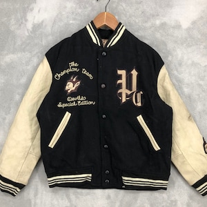Op de afbeelding: Zwart en crème varsity jack met een groot "P.C." logo op de voorkant en een "The Champion Team Devils Special Edition" patch aan de linkerkant.