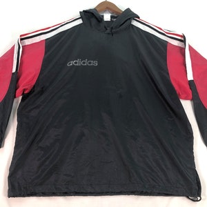 Op de afbeelding: Zwarte windjack met capuchon met rode en witte accenten. De jas heeft een ritssluiting en een capuchon met trekkoord. Het Adidas-logo is op de voorkant van de jas gedrukt.