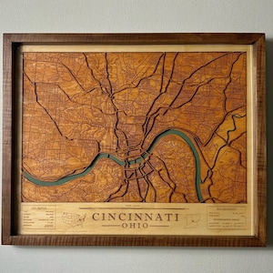 May include: A framed, three-dimensional wooden map of Cincinnati, Ohio, featuring intricate laser-cut street patterns and a teal-colored river winding through the center. The map includes text labels for city profile and map reference details.