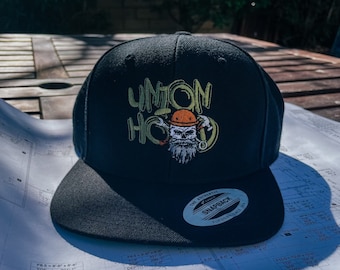 May include: A black baseball cap with a white and yellow embroidered logo that says "UNION HOOD". The logo features a cartoon image of a construction worker wearing a hard hat and holding a wrench. The cap has a black snap closure and a white sticker that says "THE AUTHENTIC SNAPBACK YUPOONS SINCE 1997".