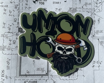 May include: A green and black sticker with the words "Union Hod" in white letters. The sticker features a cartoon skull wearing a hard hat and a beard. The sticker is placed on a blueprint with measurements in inches.