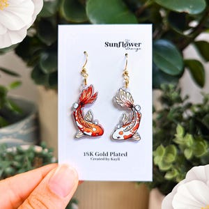 May include: A pair of gold plated earrings featuring a red and white koi fish design. The earrings are on a white card with the text "Sunflower Strange" and "18K Gold Plated Created by Kayli".