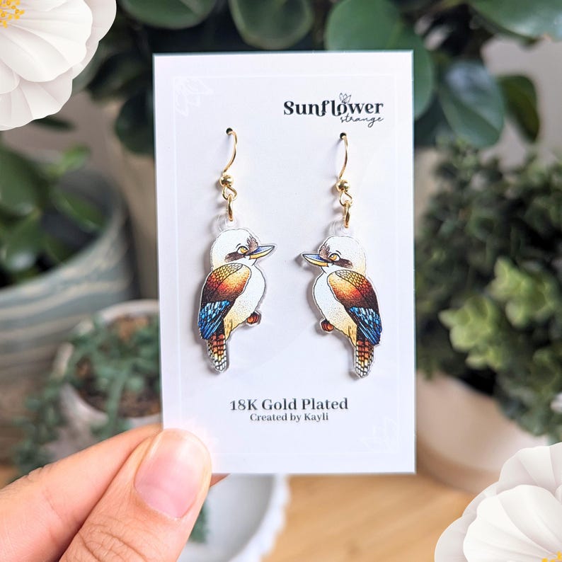 K&ouml;nnte beinhalten: Ein Paar vergoldete Ohrringe mit einem farbenfrohen Kookaburra-Motiv. Die Ohrringe befinden sich auf einer wei&szlig;en Karte mit dem Text "Sunflower Strange" und "18K Gold Plated, Created by Kayli."