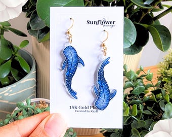 May include: A pair of gold-plated earrings featuring a blue whale shark design. The earrings are made of clear acrylic and have a gold hook closure. The earrings are labelled "18K Gold Plated" and "Created by Kayli".