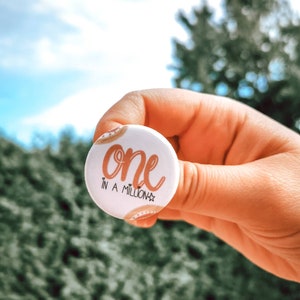 Peut inclure: Un bouton blanc avec un motif brun et or. Le bouton dit "one in a million" avec une étoile.