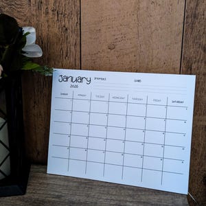 May include: A white paper calendar for January 2026, with a grid layout for daily planning. The calendar includes sections for priorities and goals. It is set on a wooden surface with a dark background.