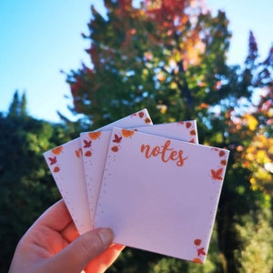 Peut inclure: Un lot de trois blocs-notes blancs avec une bordure de feuilles d'automne. Le bloc-notes du haut porte le mot "notes" écrit en cursive orange.