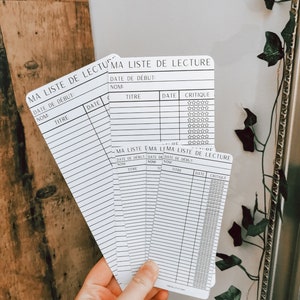 May include: A set of five white cards with black text, each labelled "Ma Liste de Lecture" (My Reading List) and featuring columns for book title, author, date started, date finished, and a rating system. The cards are designed for tracking reading progress.