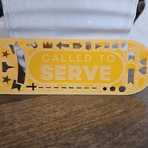 May include: A yellow rectangular sign with the words "CALLED TO SERVE" in a bold font. The sign has decorative cutouts of stars, hearts, and arrows. The sign is on a wooden surface.