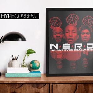 Puede incluir: P&oacute;ster enmarcado en negro con la banda N.E.R.D. con texto y gr&aacute;ficos rojos sobre fondo negro. El p&oacute;ster incluye el texto "NO ONE EVER REALLY DIES". El p&oacute;ster se muestra sobre una superficie de madera.
