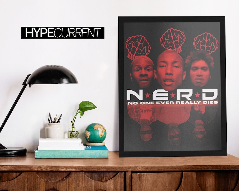 Puede incluir: P&oacute;ster enmarcado en negro con la banda N.E.R.D. con texto y gr&aacute;ficos rojos sobre fondo negro. El p&oacute;ster incluye el texto "NO ONE EVER REALLY DIES". El p&oacute;ster se muestra sobre una superficie de madera.