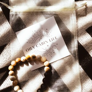 May include: A light brown and black beaded bracelet with a white card that says "Oily Cabin Life" in brown text. The card has a moon and sun graphic with rays of light.