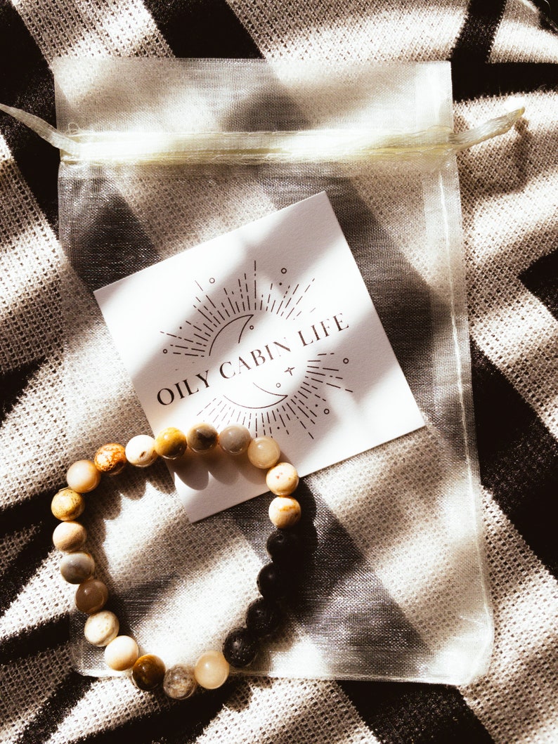 May include: A light brown and black beaded bracelet with a white card that says "Oily Cabin Life" in brown text. The card has a moon and sun graphic with rays of light.