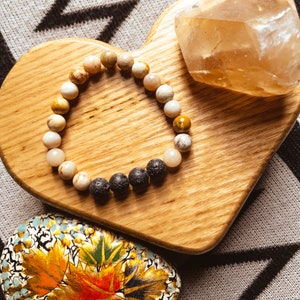 May include: A heart-shaped wooden tray with a brown and white beaded bracelet and a clear quartz crystal. The tray is on a gray and black patterned surface.