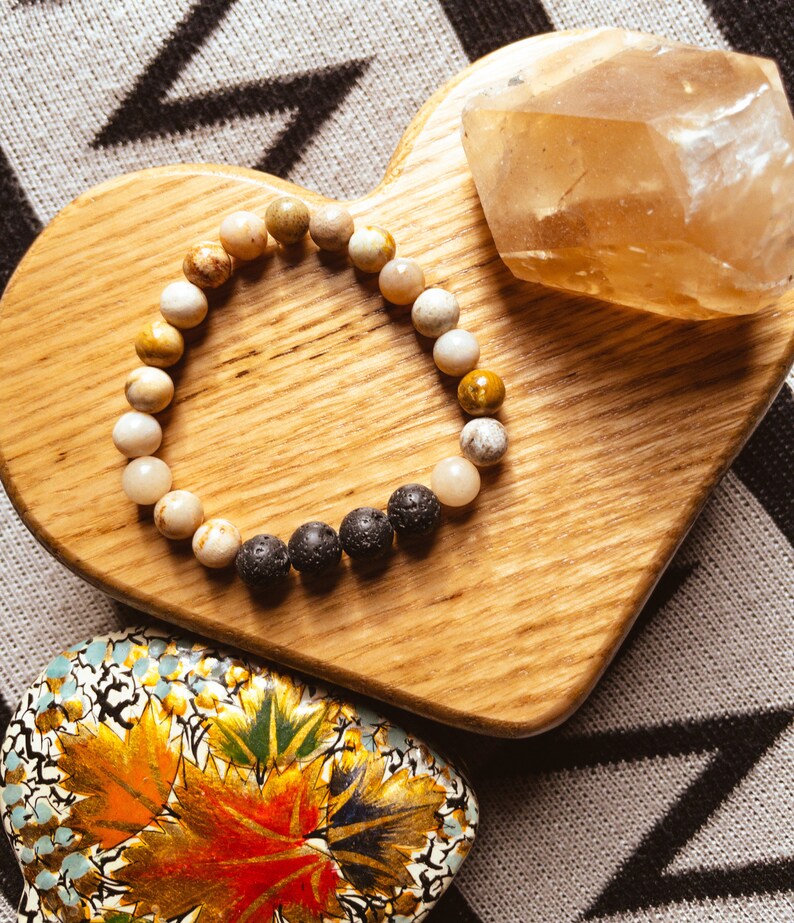 May include: A heart-shaped wooden tray with a brown and white beaded bracelet and a clear quartz crystal. The tray is on a gray and black patterned surface.