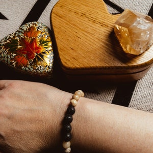May include: A black and beige beaded bracelet with lava rock beads and white agate beads. The bracelet is on a person's wrist.