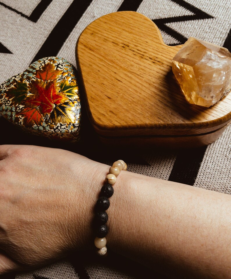 May include: A black and beige beaded bracelet with lava rock beads and white agate beads. The bracelet is on a person's wrist.