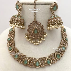 May include: An antique gold-toned jewelry set featuring a necklace, earrings, and a headpiece. The set includes teardrop-shaped, light green gemstones and small white pearls. The earrings are bell-shaped. The text "IndoExquisite Designs" is visible.