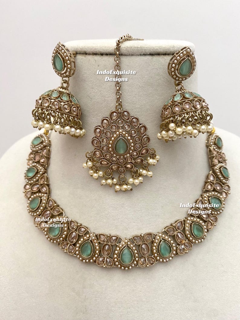 May include: An antique gold-toned jewelry set featuring a necklace, earrings, and a headpiece. The set includes teardrop-shaped, light green gemstones and small white pearls. The earrings are bell-shaped. The text "IndoExquisite Designs" is visible.