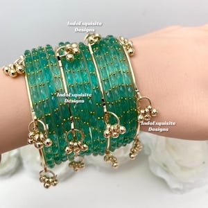May include: Emerald green bangles with gold detailing. Each bangle features small gold beads and dangling charms. The bracelets are stacked, highlighting their intricate design. The text "IndoExquisite Designs" is visible.