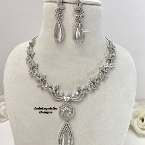 May include: A silver necklace and earrings set with a teardrop-shaped pendant and floral accents. The necklace features a delicate chain with a floral design and a large teardrop-shaped pendant with a hexagonal setting. The earrings are also teardrop-shaped with floral accents. The jewelry is made with clear cubic zirconia stones.