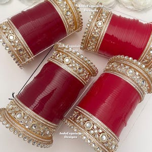 Puede incluir: Cuatro pulseras rojas con adornos de oro y cristal. Las pulseras están dispuestas en un recipiente de plástico transparente. El adorno dorado presenta intrincados diseños florales y geométricos. Hay flores blancas en el fondo.