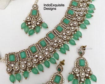 May include: A green and gold Indian bridal necklace set with a choker style necklace and matching earrings. The necklace features a row of emerald green stones and is decorated with pearls and rhinestones. The earrings are also decorated with emerald green stones, pearls, and rhinestones.