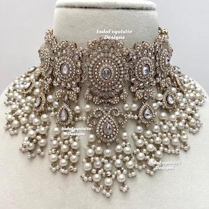 Op de afbeelding: Een sierlijke choker-ketting in antieke stijl. De ketting heeft een centraal ontwerp met heldere edelstenen en een parelfranje. Het metaal heeft een goudkleurige tint. De ketting wordt gepresenteerd op een witte ondergrond.