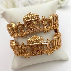 Puede incluir: Dos brazaletes de tono dorado con diseños intrincados que representan una procesión de figuras y un palanquín. Los brazaletes están apilados sobre una superficie blanca.